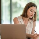 Eine junge Frau sitzt im Büro und schreibt Notizen auf einen Block neben Ihrem Laptop