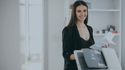 Eine Frau steht am Drucker im Büro mit gedruckten Dokumenten in der Hand und einem Lächeln auf den Lippen