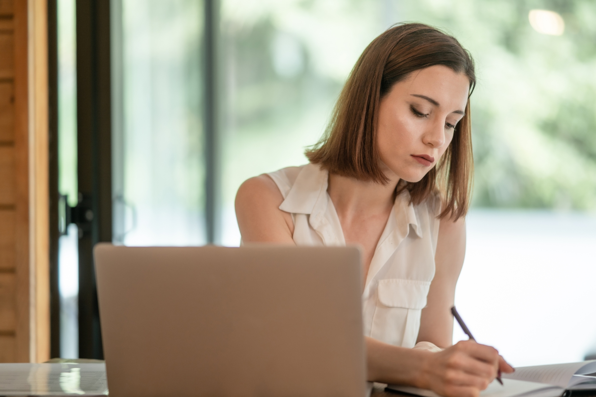 Eine junge Frau sitzt im Büro und schreibt Notizen auf einen Block neben Ihrem Laptop